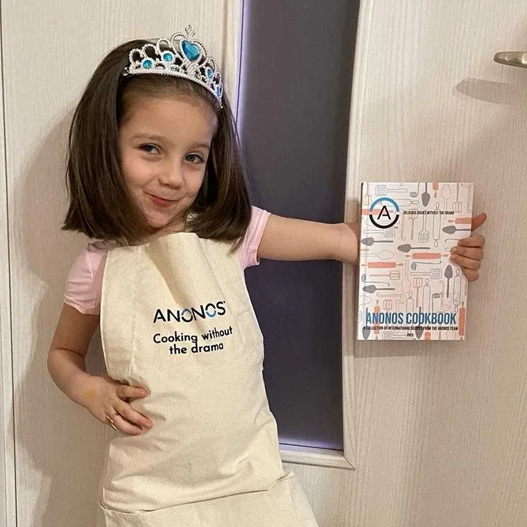 A young child wearing an Anonos 'Cooking without the drama' apron and holding the Anonos Cookbook.